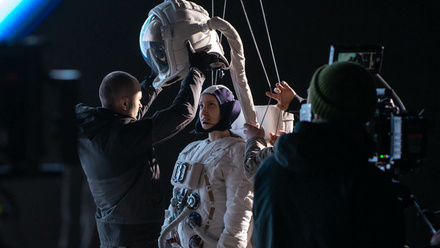 Photo on-set of a woman in an space suit with harnesses attached and a camera either side of her. Someone is helping her into her helmet.