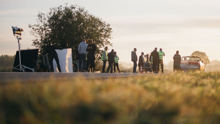 Photo taken from a low angle, a grassy field in the foreground beside a road where a film crew is set up to film a car. The sky is bright.