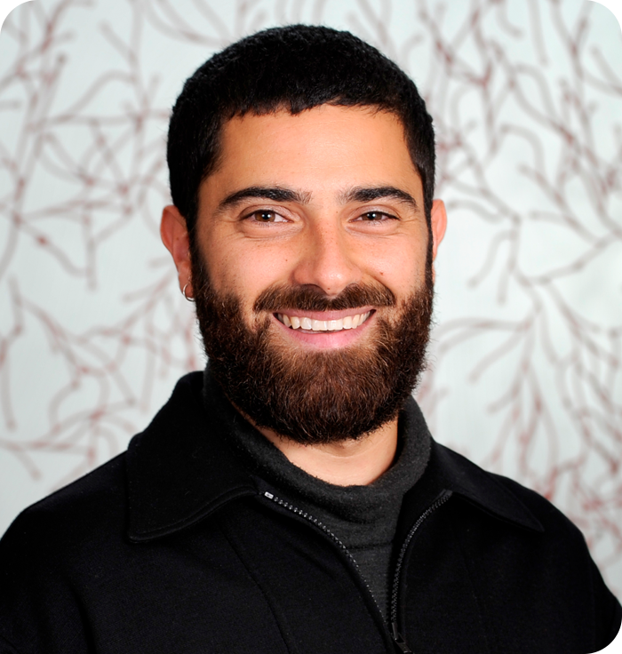 Headshot: a young man with short, black hair and a full, black beard. He is wearing a black t-shirt with a black jacket over it.