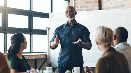 Photo of a man stood in front of a whiteboard, he is presenting to a small group of people sat around a desk directly in front of him.