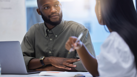 Photo of two people in a meeting, the woman is talking whilst the man listens. They have their laptop and notebooks on the desk in front of them.