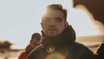 Headshot: A man with short brown hair wearing a large rain jacket, on location somewhere.