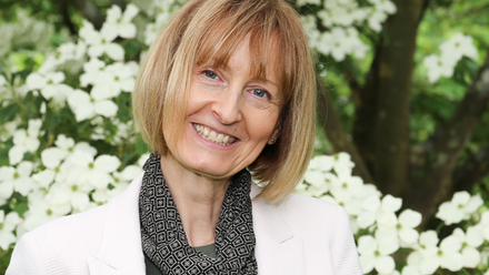 Headshot: A woman with a blonde/brown bob wearing a thin black scarf and white jacket.