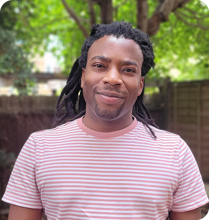 Headshot: Dean Webster, a black man with long dreadlocks tied back from his face. He is wearing a pink striped t-shirt.