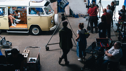 Photo taken on location, a crew of around 7 people stands by equipment whilst actors sit in a VW van waiting for filming to begin.