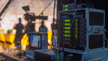 Photo on-set of a production, there is a bank of radio mics in the foreground and production crew and camera in the background.