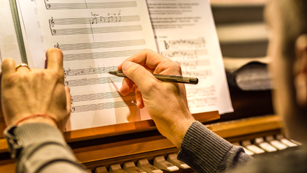 Photo close up of hands writing on a music sheet which is propped up on a piano or organ.