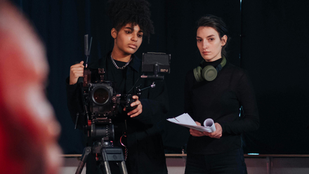 Image of two women, one is behind a camera on a tripod and the other is stood beside her with headphones on.