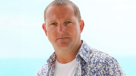 Headshot: David Notman Watt, a man with very short brown hair wearing an open patterned blue and white shirt with a white t-shirt.