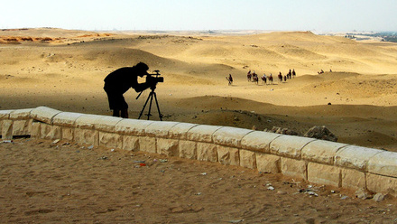 The silhouette of a cameraperson bent over a large broadcast camera on a tripod is set against a desert background with a camel train weaving through.