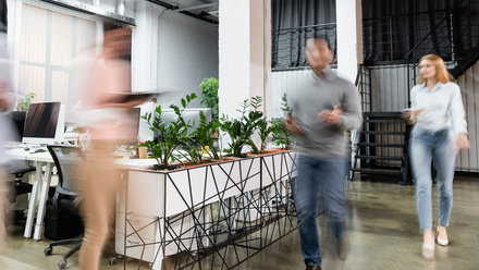 Image of a row of plants in an office space, with computers alongside them and people walking past in a blur.