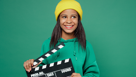 Photo image of a young girl holding a clapper board and smiling towards the camera, stood in front of a green wall.