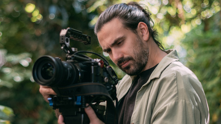 Photo of a man with long dark hair and a beard holding a broadcast camera up towards his face. He is surrounded by greenery.