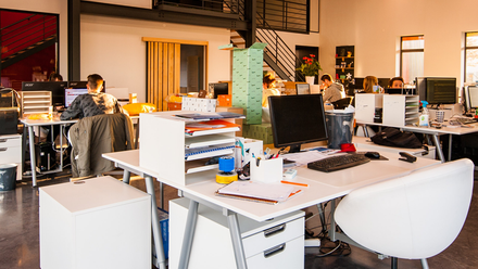 Photo of a large open-plan office space with white desks. Some people can be seen working in the background.