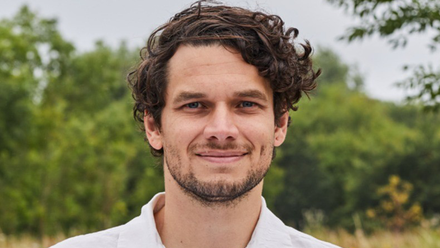 Headshot: A man with short wavy brown hair and a short beard, wearing a white shirt and standing in a field with tall grass and trees.