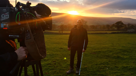 Photo of a presenter holding a white cane in a field, with the sunset behind him. There is a camera on a tripod on the left of shot, pointing at him.