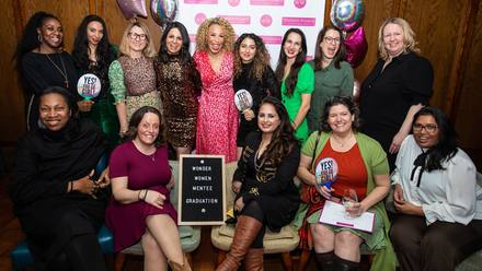 Photo image of a diverse group of women, smiling widely at the camera with some balloons and a step and repeat board in the background.