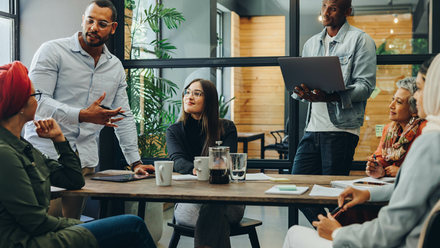 Photo of a diverse team at work, sat and stood around a table brainstorming ideas.