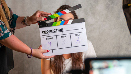 Photo of a clapper board being held up in front of a woman with long brown hair who is sat in front of a grey background. A monitor is just visible.