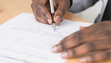 Photo close up of somebody signing a contract, only the desk, contract, hands and pen are visible.