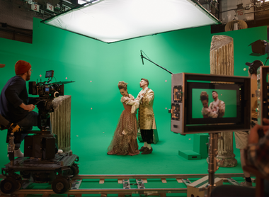 Photo of a greenscreen studio, two actors in period costumes are stood in the middle of the stage. There is a camera on a track being and monitors.