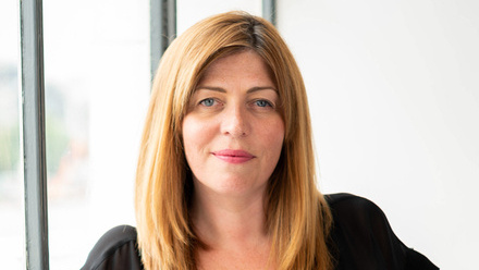 Photo of a woman with long, straight light brown hair wearing a black top, stood in front of a large window.