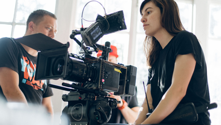 Photo of a woman sat behind a film camera, in a large light space.