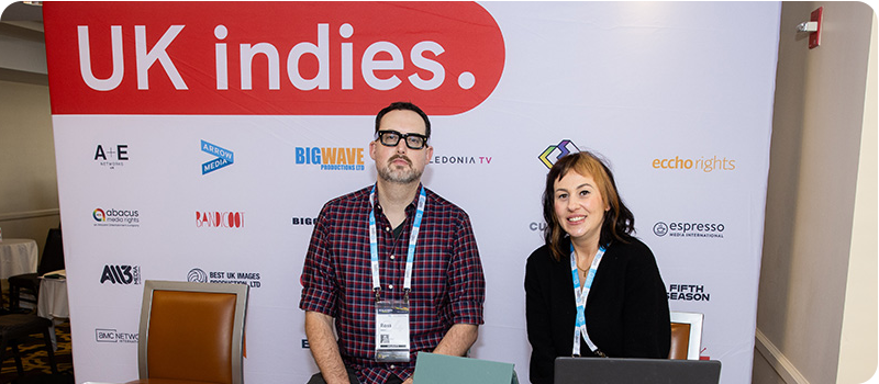 Photo of Pact's Ross Lewis and Aimee Shimmin sitting behind a desk surrounded by UK Indies branding at the 2024 Realscreen Summit.