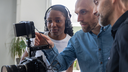 Photo of a small crew stood behind a camera, looking at the screen. A female member of the crew wears headphones.