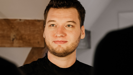 Headshot: A young man with short brown hair and a short brown beard wearing a black top.