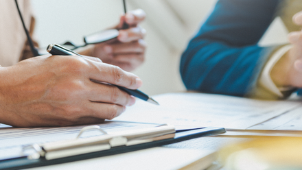 Photo close up of two mens' hands on a desk with papers in front of them. One is holding a pen over one of the documents.