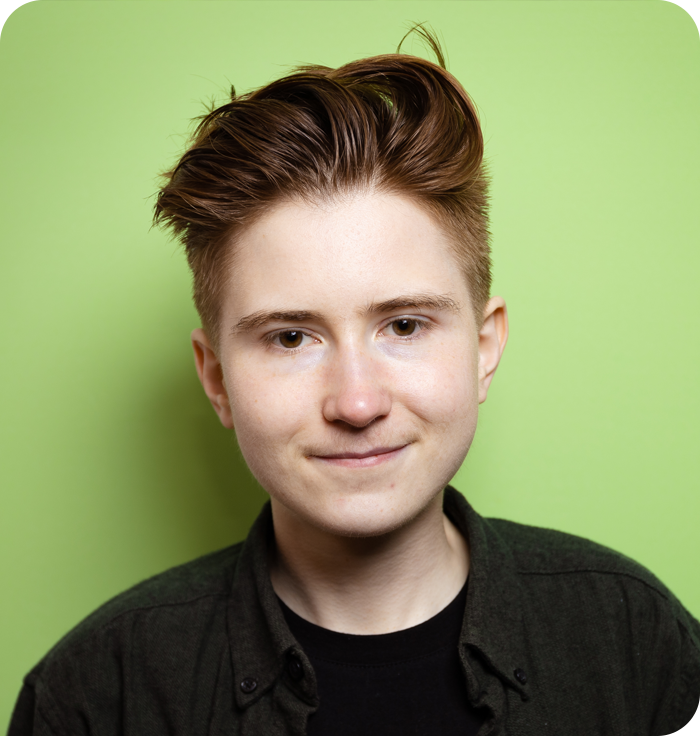 Headshot: a young man with quaffed red hair. He is wearing a black t-shirt and black shirt over the top of it.