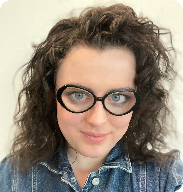 Headshot: Darcy Dear - a woman with curly brown, shoulder-length hair wearing dark-rimmed glasses and a denim shirt.