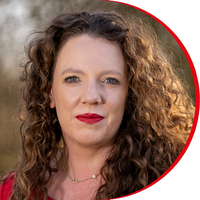 Headshot: Kate Beal, a woman with long curly brown hair, she is wearing red lipstick and a red top.