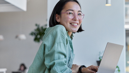 Image of a desktop with an open laptop and phone beside it, a woman wearing a pastel green shirt and glasses is sat beside it.