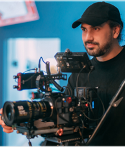 Photo of a camera person stood behind a broadcast camera on a tripod. They are facing forwards and look engaged.