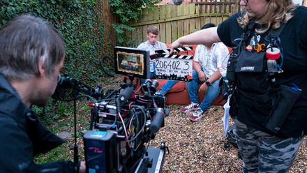 Photo of a camera set up in a small courtyard garden, pointing at two actors sitting on a sofa. A woman holds a clapper board in front of the camera.