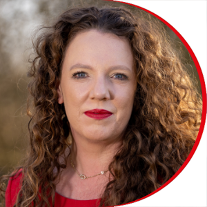 Headshot: Kate Beal - a woman with long curly brown hair, she is wearing red lipstick and a red top.