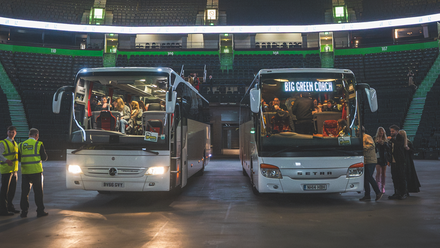 Photo of two white coaches parked inside an empty arena with some people in high vis and passengers stood beside them.