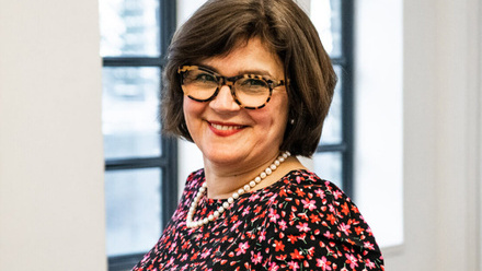 Headshot: Ulla Streib, a woman with a bobbed brown haircut wearing statement glasses and a flower-patterned dress.