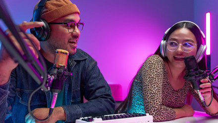 Photo of two podcasters sat behind a desk with microphones in front of them, engaged in a recording. Atmospheric purple and blue lighting behind them.