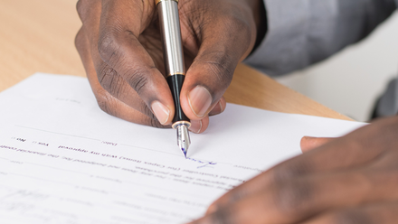 Photo close up of a pair of hands. The right hand is holding a pen and signing a contract on a desk.