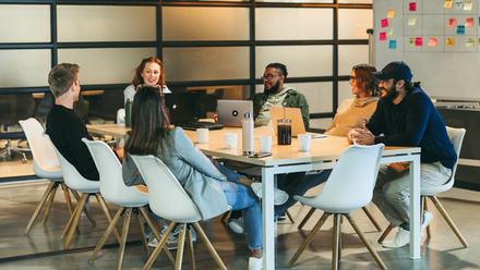 Photo of a diverse team sat around a meeting table in a light office space with a white board full of colourful post-it notes behind them.