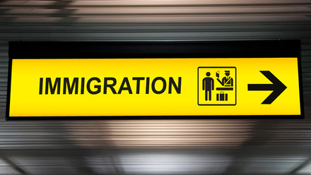 Photo of a bright yellow ceiling sign with the word 'IMMIGRATION' in black, a small icon image of an immigration desk and an arrow pointing right.