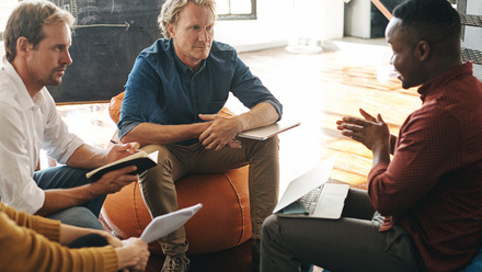 Photo of a diverse group of three men sat in a meeting in an informal part of the office, with notebooks in front of them.