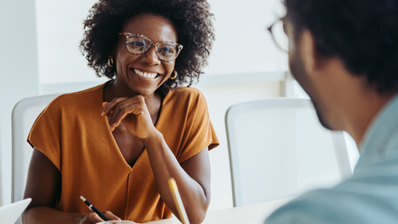 Photo of a woman having a meeting with a man opposite her, she is smiling and engaged and they both have pens and notebooks.