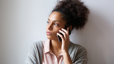 Photo of a woman on her mobile, stood against a wall. She is looking off into the distance.