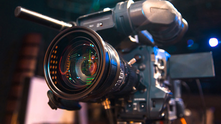 Photo image of a broadcast camera, quite close up, in a dark studio with just a few spotlights behind the camera.