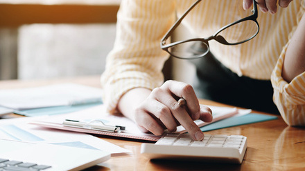 Photo close up of a calculator on a desk. Someone is leaning over it and pressing a button whilst holding their glasses in their other hand.