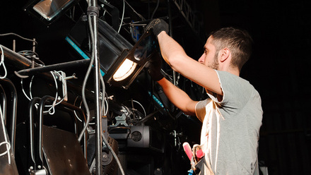 Photo of a crew member wearing a tool belt and gloves adjusting a light on a rig.